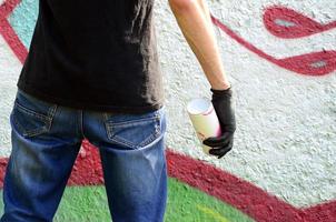 A young hooligan with a spray can stands against a concrete wall with graffiti paintings. Illegal vandalism concept. Street art photo