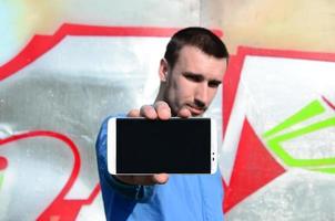 The graffiti artist demonstrates a smartphone with an empty black screen against the background of a colorful painted wall. Street art concept photo