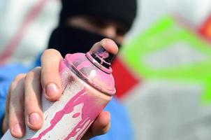 A young graffiti artist in a blue jacket and black mask is holding a can of paint in front of him against a background of colored graffiti drawing. Street art and vandalism concept photo