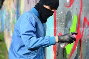 A young hooligan with a hidden face paints graffiti on a metal wall. Illegal vandalism concept photo