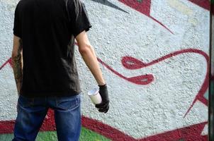 A young hooligan with a spray can stands against a concrete wall with graffiti paintings. Illegal vandalism concept. Street art photo