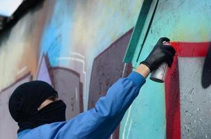 A young hooligan with a hidden face paints graffiti on a metal wall. Illegal vandalism concept photo