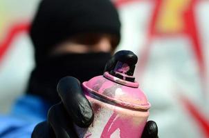 A young graffiti artist in a blue jacket and black mask is holding a can of paint in front of him against a background of colored graffiti drawing. Street art and vandalism concept photo