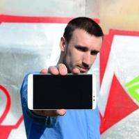 The graffiti artist demonstrates a smartphone with an empty black screen against the background of a colorful painted wall. Street art concept photo