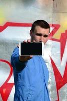 The graffiti artist demonstrates a smartphone with an empty black screen against the background of a colorful painted wall. Street art concept photo