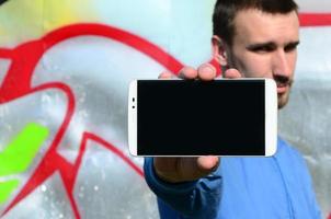 The graffiti artist demonstrates a smartphone with an empty black screen against the background of a colorful painted wall. Street art concept photo