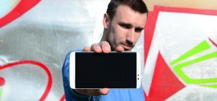 The graffiti artist demonstrates a smartphone with an empty black screen against the background of a colorful painted wall. Street art concept photo