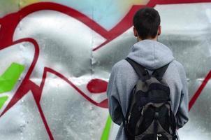 A young graffiti artist with a black bag looks at the wall with his graffiti on a wall. Street art concept photo