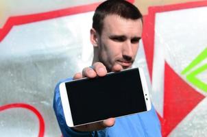 The graffiti artist demonstrates a smartphone with an empty black screen against the background of a colorful painted wall. Street art concept photo