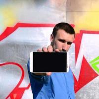 el artista de graffiti muestra un teléfono inteligente con una pantalla negra vacía contra el fondo de una pared pintada de colores. concepto de arte callejero foto