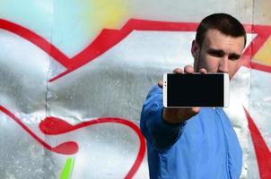 The graffiti artist demonstrates a smartphone with an empty black screen against the background of a colorful painted wall. Street art concept photo