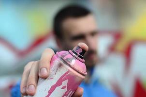 un joven artista de graffiti con una chaqueta azul sostiene una lata de pintura frente a él sobre un fondo de dibujo de graffiti de colores. concepto de arte callejero y vandalismo foto