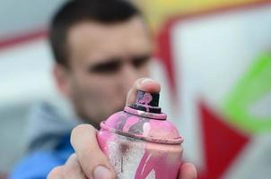 un joven artista de graffiti con una chaqueta azul sostiene una lata de pintura frente a él sobre un fondo de dibujo de graffiti de colores. concepto de arte callejero y vandalismo foto