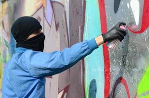 A young hooligan with a hidden face paints graffiti on a metal wall. Illegal vandalism concept photo