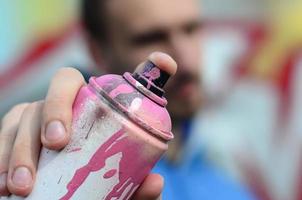 A young graffiti artist in a blue jacket is holding a can of paint in front of him against a background of colored graffiti drawing. Street art and vandalism concept photo