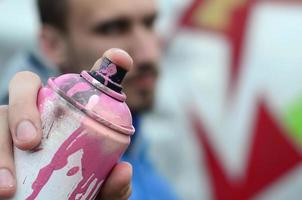 A young graffiti artist in a blue jacket is holding a can of paint in front of him against a background of colored graffiti drawing. Street art and vandalism concept photo