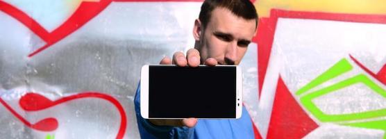 The graffiti artist demonstrates a smartphone with an empty black screen against the background of a colorful painted wall. Street art concept photo