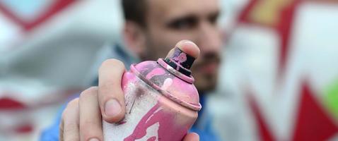 A young graffiti artist in a blue jacket is holding a can of paint in front of him against a background of colored graffiti drawing. Street art and vandalism concept photo