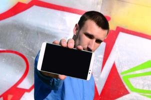 el artista de graffiti muestra un teléfono inteligente con una pantalla negra vacía contra el fondo de una pared pintada de colores. concepto de arte callejero foto