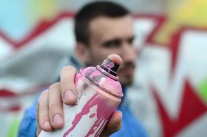 A young graffiti artist in a blue jacket is holding a can of paint in front of him against a background of colored graffiti drawing. Street art and vandalism concept photo