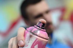 un joven artista de graffiti con una chaqueta azul sostiene una lata de pintura frente a él sobre un fondo de dibujo de graffiti de colores. concepto de arte callejero y vandalismo foto