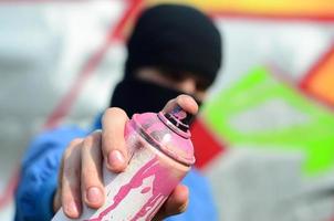 un joven artista de graffiti con una chaqueta azul y una máscara negra sostiene una lata de pintura frente a él contra un fondo de dibujo de graffiti de colores. concepto de arte callejero y vandalismo foto