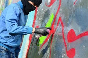 un joven hooligan con un rostro oculto pinta graffiti en una pared de metal. concepto de vandalismo ilegal foto