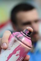 A young graffiti artist in a blue jacket is holding a can of paint in front of him against a background of colored graffiti drawing. Street art and vandalism concept photo