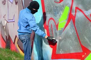 un joven hooligan con un rostro oculto pinta graffiti en una pared de metal. concepto de vandalismo ilegal foto
