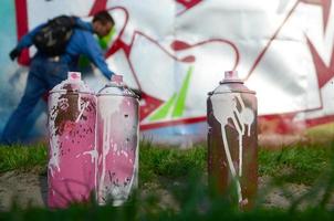 A few used paint cans against the background of the space with the wall on which the young guy draws a large graffiti drawing. Modern art of drawing walls in graffiti photo