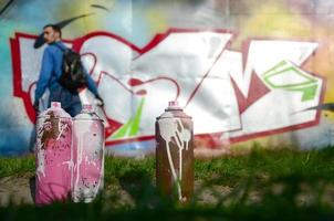 A few used paint cans against the background of the space with the wall on which the young guy draws a large graffiti drawing. Modern art of drawing walls in graffiti photo