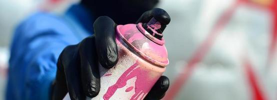 A young graffiti artist in a blue jacket and black mask is holding a can of paint in front of him against a background of colored graffiti drawing. Street art and vandalism concept photo