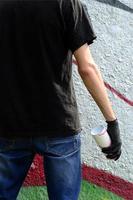 A young hooligan with a spray can stands against a concrete wall with graffiti paintings. Illegal vandalism concept. Street art photo