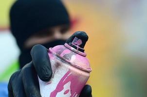 A young graffiti artist in a blue jacket and black mask is holding a can of paint in front of him against a background of colored graffiti drawing. Street art and vandalism concept photo