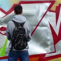 A young graffiti artist with a black bag looks at the wall with his graffiti on a wall. Street art concept photo