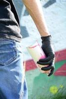 A young hooligan with a spray can stands against a concrete wall with graffiti paintings. Illegal vandalism concept. Street art photo