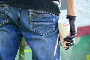 un joven hooligan con una lata de aerosol se para contra una pared de hormigón con pinturas de graffiti. concepto de vandalismo ilegal. arte callejero foto