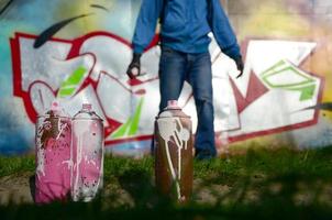 A few used paint cans against the background of the space with the wall on which the young guy draws a large graffiti drawing. Modern art of drawing walls in graffiti photo