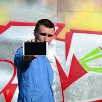 The graffiti artist demonstrates a smartphone with an empty black screen against the background of a colorful painted wall. Street art concept photo