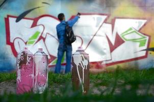 unas cuantas latas de pintura usadas contra el fondo del espacio con la pared en la que el joven dibuja un gran dibujo de graffiti. arte moderno de dibujar paredes en graffiti foto