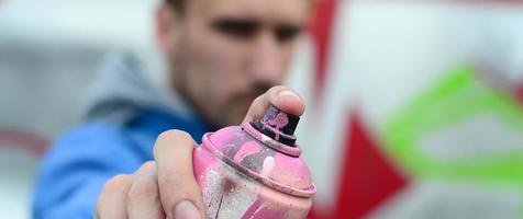 A young graffiti artist in a blue jacket is holding a can of paint in front of him against a background of colored graffiti drawing. Street art and vandalism concept photo