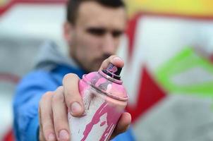 A young graffiti artist in a blue jacket is holding a can of paint in front of him against a background of colored graffiti drawing. Street art and vandalism concept photo