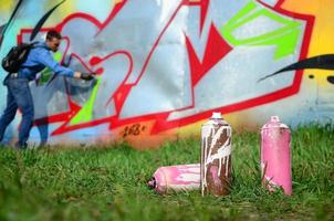 A few used paint cans against the background of the space with the wall on which the young guy draws a large graffiti drawing. Modern art of drawing walls in graffiti photo