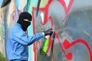 A young hooligan with a hidden face paints graffiti on a metal wall. Illegal vandalism concept photo