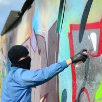 A young hooligan with a hidden face paints graffiti on a metal wall. Illegal vandalism concept photo