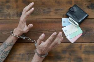 manos esposadas de sospechoso criminal tatuado de cardado y tarjetas de crédito falsas con dinero en efectivo en el monedero como evidencia para la investigación foto