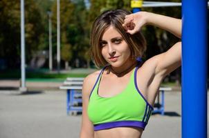 retrato de una joven atleta caucásica con ropa deportiva verde brillante en el contexto de un gimnasio deportivo callejero para entrenamiento de atletismo al aire libre. deportes de verano al aire libre foto