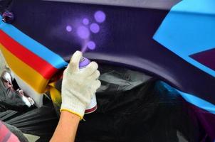 A hand with a spray can that draws a new colorful graffiti on the car. Photo of the process of drawing a graffiti on an automobile close-up. The concept of street art and illegal vandalism