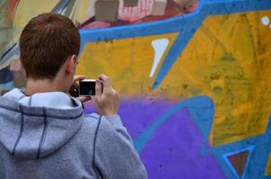 un joven grafitero fotografía su cuadro completo en la pared. el chico usa tecnología moderna para capturar un colorido dibujo de graffiti abstracto. centrarse en el dispositivo de fotografía foto