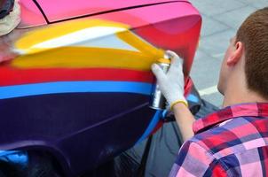 A young red-haired graffiti artist paints a new colorful graffiti on the car. Photo of the process of drawing a graffiti on a car close-up. The concept of street art and illegal vandalism